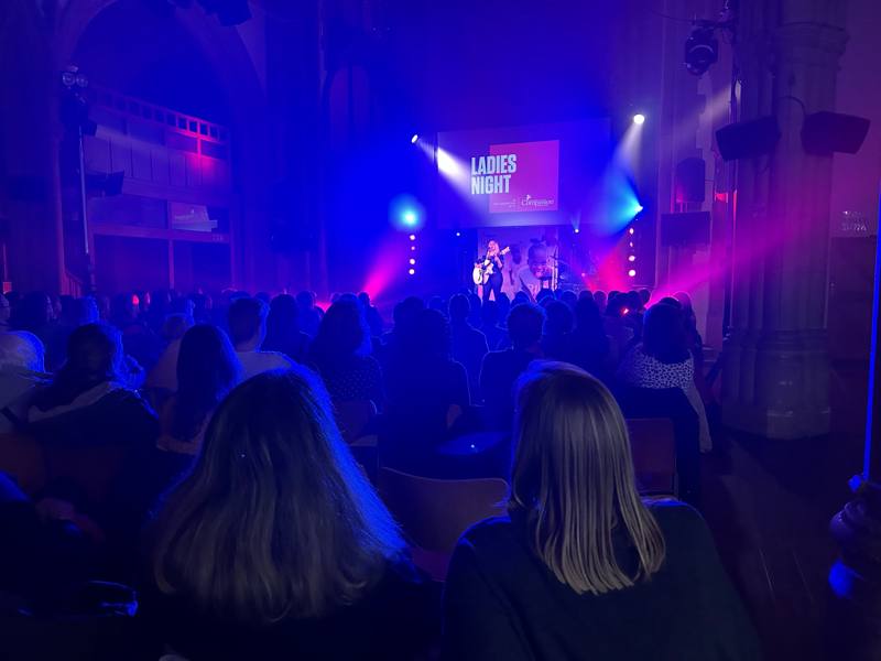 Ladies Night tour: a singer with a guitar on a church stage under blue and pink lights, the Ladies Night title on the screen, a full seated audience in front